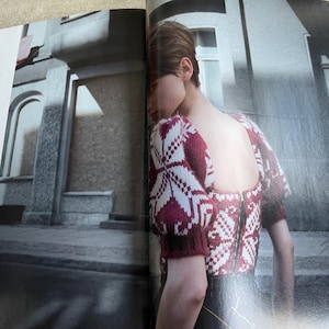 May include: A burgundy and white patterned sweater with a zipper back. The sweater has short sleeves and a square neckline. The image is from a fashion magazine, with a blurred background of a building.