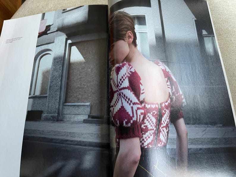 May include: A burgundy and white patterned sweater with a zipper back. The sweater has short sleeves and a square neckline. The image is from a fashion magazine, with a blurred background of a building.