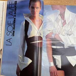 May include: Fashion magazine spread featuring two models. One model wears a white collared shirt with a black vest and skirt. The other model wears a white shirt with diamond-shaped embroidery and black shorts. The text "LA SOIE LAVEE UNE MATIERE A SCULPTER" is visible.