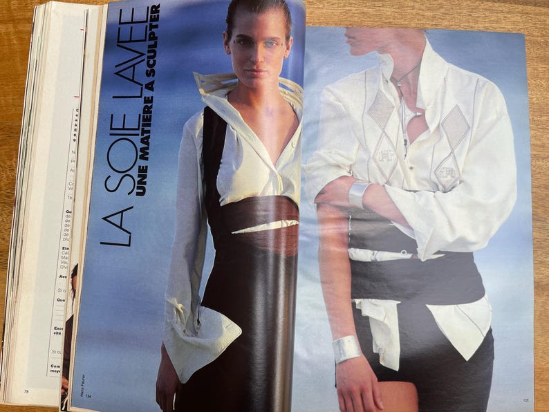 May include: Fashion magazine spread featuring two models. One model wears a white collared shirt with a black vest and skirt. The other model wears a white shirt with diamond-shaped embroidery and black shorts. The text "LA SOIE LAVEE UNE MATIERE A SCULPTER" is visible.