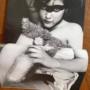 May include: Black and white photo of a woman wearing a black eye mask, holding a stuffed animal. The photo is from a magazine and has the text "Photographed by Louis D'Amico" in the top right corner.