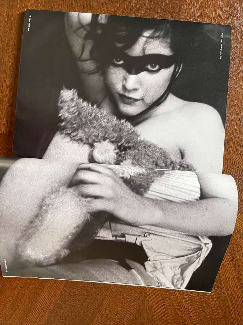 May include: Black and white photo of a woman wearing a black eye mask, holding a stuffed animal. The photo is from a magazine and has the text "Photographed by Louis D'Amico" in the top right corner.
