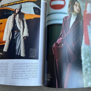 Puede incluir: Una revista de moda de doble p&aacute;gina. Una modelo lleva un abrigo largo de piel sint&eacute;tica color crema, pantalones grises y un gorro gris. La otra modelo lleva un traje de pana burdeos con un cuello alto blanco. El fondo incluye un veh&iacute;culo amarillo y una se&ntilde;al roja y blanca.