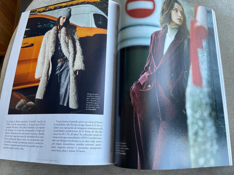 Puede incluir: Una revista de moda de doble p&aacute;gina. Una modelo lleva un abrigo largo de piel sint&eacute;tica color crema, pantalones grises y un gorro gris. La otra modelo lleva un traje de pana burdeos con un cuello alto blanco. El fondo incluye un veh&iacute;culo amarillo y una se&ntilde;al roja y blanca.