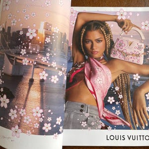 Puede incluir: Anuncio de revista con una mujer que lleva una bufanda rosa y sostiene un bolso rosa con detalles florales. El fondo muestra un paisaje urbano con ilustraciones de flores de cerezo y el texto "LOUIS VUITTON".