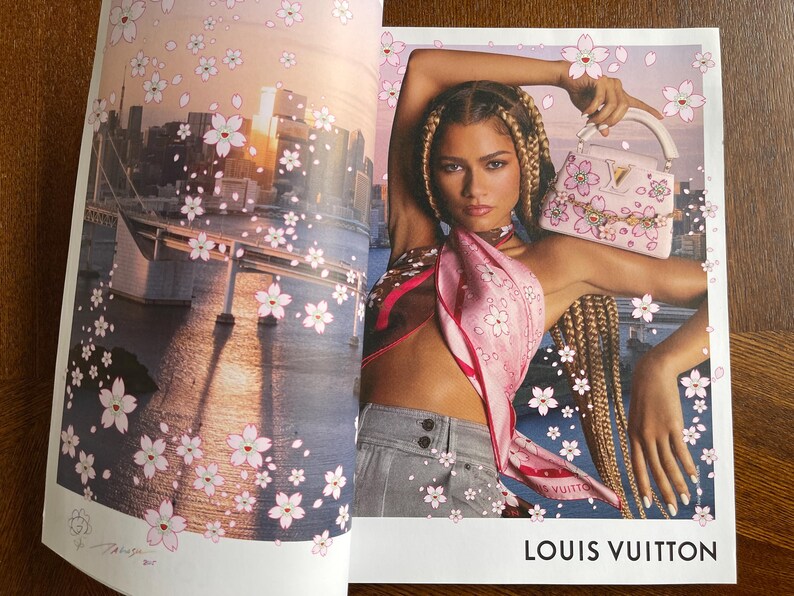 Puede incluir: Anuncio de revista con una mujer que lleva una bufanda rosa y sostiene un bolso rosa con detalles florales. El fondo muestra un paisaje urbano con ilustraciones de flores de cerezo y el texto "LOUIS VUITTON".