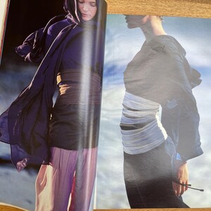 May include: A fashion magazine spread featuring two models. One model wears a dark purple draped garment with a matching head covering, a brown belt, and light purple pants. The other model wears a dark outfit with a white sash.
