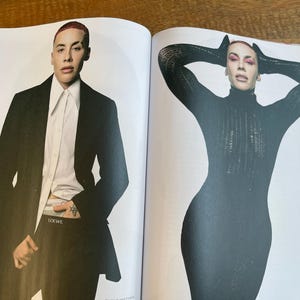May include: Two-page magazine spread featuring a person in a black suit and a person in a black turtleneck dress. The person on the left is wearing a black blazer over a white shirt and black pants. The person on the right is wearing a black turtleneck dress.