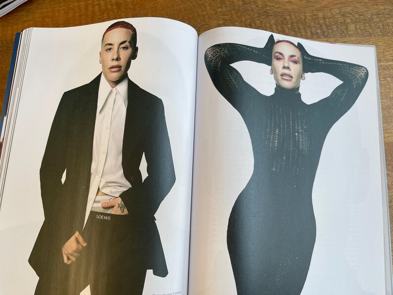 May include: Two-page magazine spread featuring a person in a black suit and a person in a black turtleneck dress. The person on the left is wearing a black blazer over a white shirt and black pants. The person on the right is wearing a black turtleneck dress.