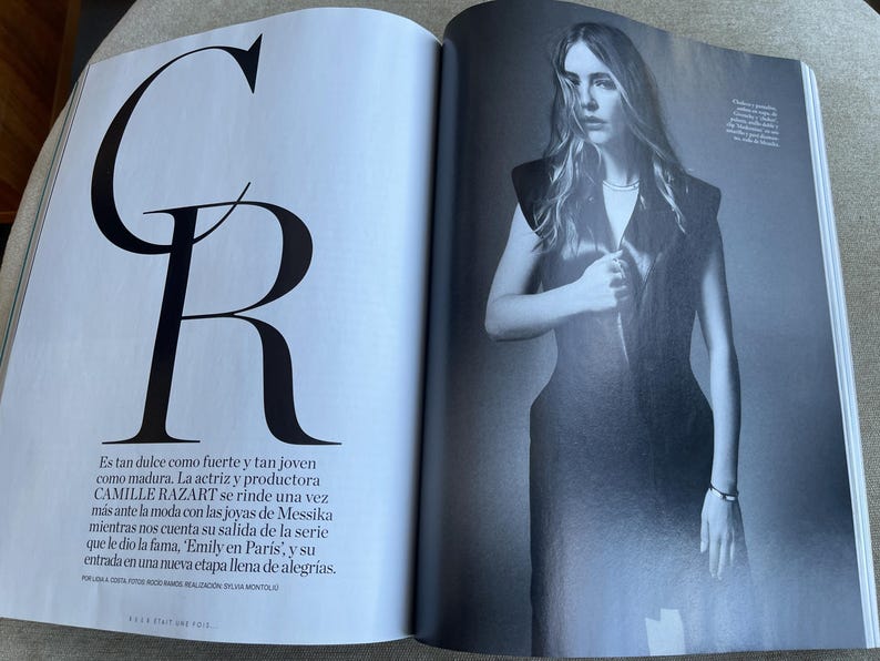 Puede incluir: Una revista con una fotograf&iacute;a en blanco y negro de una mujer con un vestido oscuro. La p&aacute;gina izquierda muestra las letras grandes "C" y "R" en negro. Texto en espa&ntilde;ol que describe a Camille Razart.
