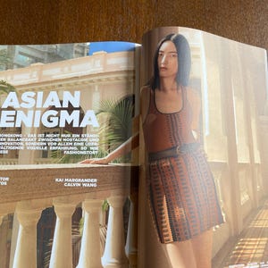 Puede incluir: Una p&aacute;gina de revista de moda con el titular "ASIAN ENIGMA". La imagen muestra a una modelo con un atuendo estampado en marr&oacute;n y naranja, de pie cerca de un balc&oacute;n. El texto incluye los nombres del editor y el fot&oacute;grafo.
