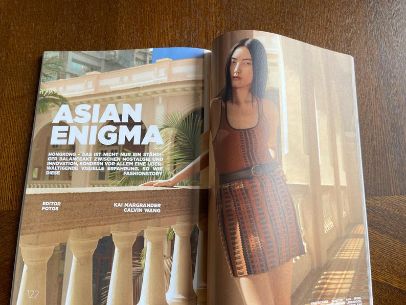 Puede incluir: Una p&aacute;gina de revista de moda con el titular "ASIAN ENIGMA". La imagen muestra a una modelo con un atuendo estampado en marr&oacute;n y naranja, de pie cerca de un balc&oacute;n. El texto incluye los nombres del editor y el fot&oacute;grafo.