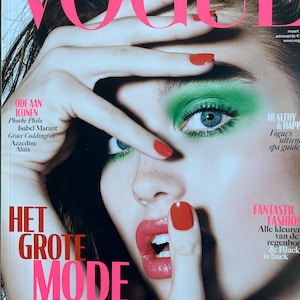 Puede incluir: Una portada de la revista Vogue Nederland con una modelo con sombra de ojos verde brillante, pintalabios rojo y esmalte de uñas rojo. La portada tiene el texto "VOGUE" en letras rosas grandes y las palabras "HET GROTE MODE NUMMER".