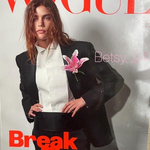 May include: Vogue Italia magazine cover. A person in a black suit, white shirt, and a pink flower is featured. The cover text reads "VOGUE ITALIA" in red, with "Break The Dress Code" in bold red letters.