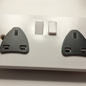 Puede incluir: Una tapa de toma de corriente blanca con dos tapas de seguridad de plástico negro. La tapa de la toma de corriente tiene dos interruptores.