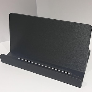 Puede incluir: Un soporte para teléfono negro de madera. El soporte tiene una parte posterior elevada y una ranura para sujetar un teléfono.