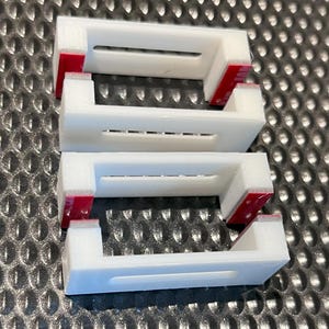 Puede incluir: Tres soportes de pared de plástico blanco con tiras adhesivas rojas en la parte posterior. Los soportes tienen una forma rectangular con una ranura en el centro.