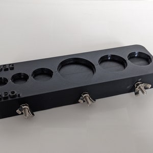 Puede incluir: Calibrador de anillos de plástico negro con cinco anillos de diferentes tamaños y tres tuercas de mariposa plateadas. El calibrador está etiquetado como "ZERO" y "10.5".