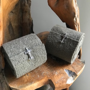 Peut inclure: Deux articles en feutre gris roulés avec des fermoirs décoratifs en argent. Le feutre est de couleur gris-vert sourd. Les fermoirs sont ornés, avec un motif en forme de croix. Les articles sont présentés sur une surface en bois rustique.