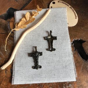 Puede incluir: Dos colgantes con forma de cruz de color bronce sobre una tela texturizada de color gris claro. También hay una ramita y una ramita de hojas secas. Una pequeña etiqueta blanquecina con el texto "Créations Artisanales" está en el fondo.