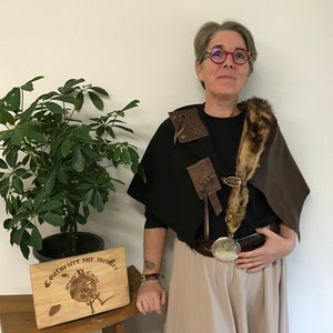 Puede incluir: Una persona lleva una capa de cuero y piel marrón con una camisa negra y un cinturón marrón con una hebilla plateada grande. La persona está de pie frente a una pared blanca con una planta en maceta a la izquierda. Un letrero de madera con el texto "Couturiere sur mesure" está sobre una mesa frente a la planta.