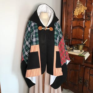 Könnte beinhalten: Ein schwarzer Kapuzenumhang mit Patchwork-Design aus grünem, rotem und weißem kariertem Stoff. Der Umhang hat einen Knebelverschluss und ist mit weißem Stoff gefüttert.