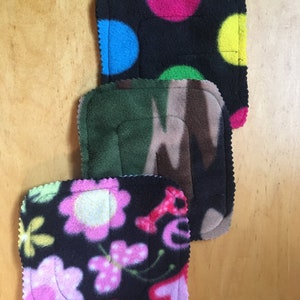 May include: Three black fleece squares with different patterns. The top square has pink, blue, green, and yellow polka dots. The middle square has a camouflage pattern. The bottom square has pink flowers, butterflies, and hearts.