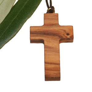 May include: A wooden cross pendant with a brown olive wood finish, hanging from a brown cord. The cross is surrounded by green olive leaves.