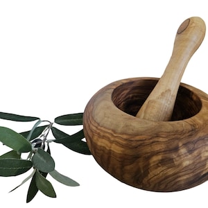 Könnte beinhalten: Ein Mörser und Stößel-Set aus Olivenholz. Der Mörser ist dunkelbraun mit einem natürlichen Holzmaserungsmuster. Der Stößel ist hellbraun und hat einen glatten, abgerundeten Griff.