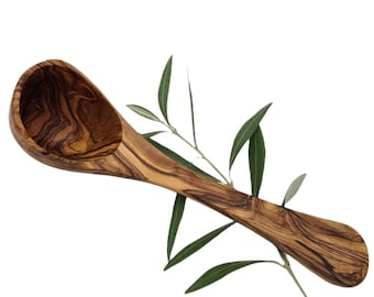 May include: A wooden olive wood ladle with a natural wood grain pattern. The ladle is brown and has a long handle. The ladle is resting on a branch of olive leaves.