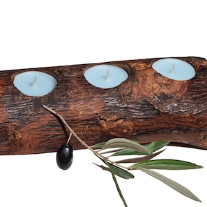 Peut inclure: Un bougeoir en bois rustique avec trois bougeoirs pour bougies chauffe-plat. Le bougeoir est fait d'un morceau de bois naturel avec une texture rugueuse, semblable à de l'écorce. Une branche d'olivier avec des feuilles vertes et une olive noire est placée devant le bougeoir.