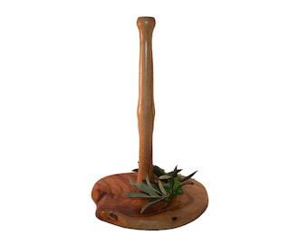 May include: A wooden paper towel holder with a natural wood finish. The holder has a round base and a tall, slender pole. A sprig of greenery with small green olives is placed on the base.