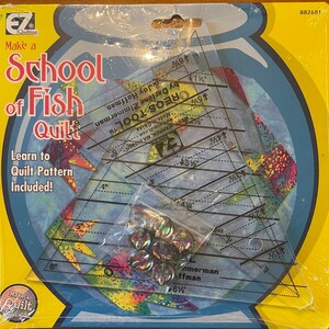 May include: A yellow and blue packaging for a quilting kit with a clear plastic ruler and a bag of iridescent beads. The packaging has the text "Make a School of Fish Quilt" and "Learn to Quilt Pattern Included!"