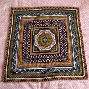 May include: A square crochet blanket with a multi-colored geometric pattern in shades of brown, green, pink, and white. The center of the blanket features a large, intricate floral design.