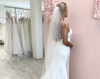 May include: A white wedding dress with a long train and a simple, elegant design. The dress has a fitted bodice and a flowing skirt. The bride is wearing a white veil.