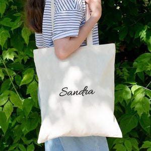 Puede incluir: Una bolsa de lona blanca con el nombre "Sandra" impreso en letra cursiva negra.