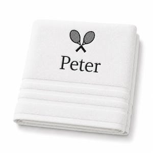 Peut inclure: Une serviette en coton blanc pliée avec une broderie représentant deux raquettes de tennis noires croisées au-dessus du prénom Peter écrit en police serif noire. La serviette présente trois bandes décoratives horizontales en bas.