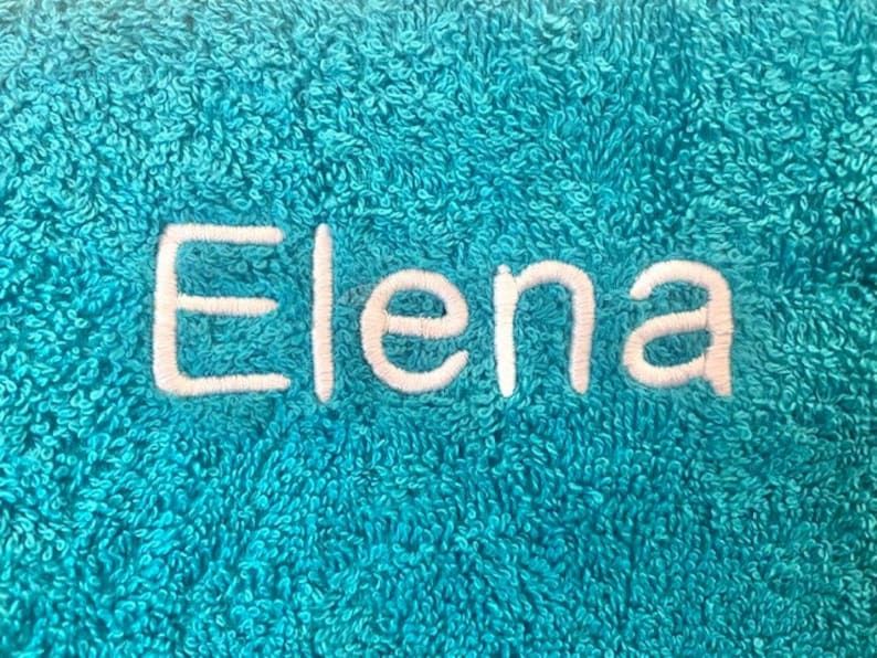 Pu&ograve; includere: Un asciugamano blu turchese con il nome "Elena" ricamato in filo bianco.