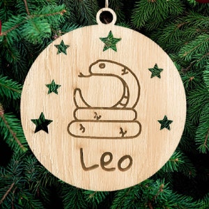 Puede incluir: Un adorno de Navidad de madera con un diseño de serpiente y la palabra "Leo" inscrita en él. El adorno está decorado con estrellas.