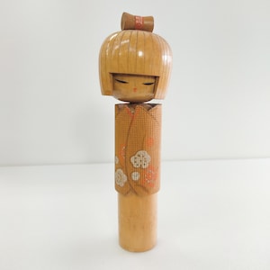 Könnte beinhalten: Japanische Kokeshi-Puppe aus Holz mit rundem Kopf und zylindrischem Körper. Die Puppe hat eine hellbraune Oberfläche mit einer kleinen Schleife auf dem Kopf. Der Körper ist mit weißen Wolken- und orangefarbenen Blumenmustern verziert.