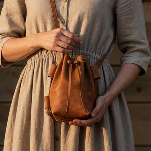 May include: A woman wearing a beige linen dress and holding a small, brown leather bucket bag with a drawstring closure and a long shoulder strap. The bag is a warm, earthy tone, and the dress has a simple, elegant design.
