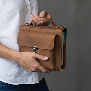 May include: A brown leather briefcase with a flap closure and a top handle. The briefcase features a front pocket and a silver metal clasp. The leather has a distressed appearance. The briefcase is designed for carrying documents and other items.