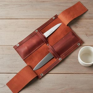 May include: A brown leather knife roll with two stainless steel knives secured inside. The roll is open, revealing the knives, and is next to a white ceramic mug. The roll is on a wooden surface.