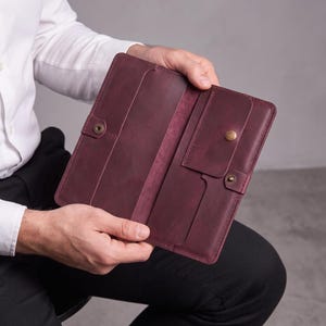 Könnte beinhalten: Ein langes, rechteckiges Portemonnaie aus dunkelviolettem Leder, das geöffnet ist und sein Inneres zeigt. Es verfügt über mehrere Kartenfächer, ein Mittelfach und eine kleine Klappentasche mit einem messingfarbenen Druckknopf.