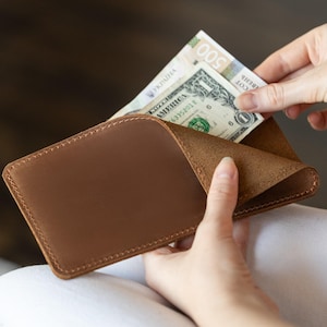 May include: A brown leather wallet with a curved top edge. The wallet is open, revealing a one-dollar bill and a Ukrainian 500-hryvnia note. The wallet is held open by a person's hands.