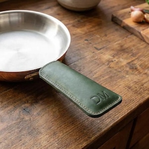 May include: A copper-bottomed stainless steel frying pan with a green leather handle cover. The handle cover is embossed with the initials "DM". The pan sits on a wooden surface, with other kitchen items in the background.
