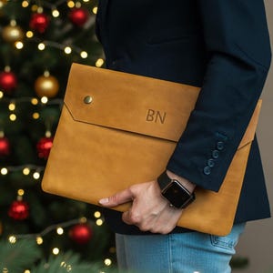 May include: A tan leather laptop sleeve with a flap closure and a brass snap. The initials "BN" are embossed on the front. The person is wearing a navy blazer and blue jeans. A Christmas tree with red and gold ornaments is in the background.
