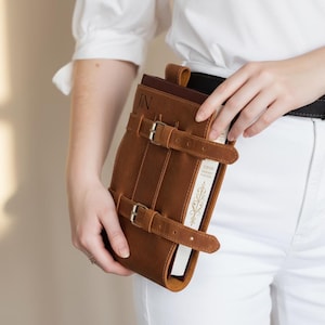 Handmade from premium leather, this medieval-inspired leather book belt holder keeps your favorite book secure and hands-free. Attaching easily to most belts, it fits journals, notebooks, and small books. Ideal for festivals, travel, cosplay, or daily use. Personalization available to create a unique custom book holder for any reader or fantasy enthusiast.