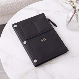 Pode incluir: Uma pasta retangular preta com fecho de correr lateral, três rebites metálicos prateados ao longo da borda esquerda e dois bolsos exteriores. O bolso inferior tem gravadas as letras douradas KD numa fonte com serifa.