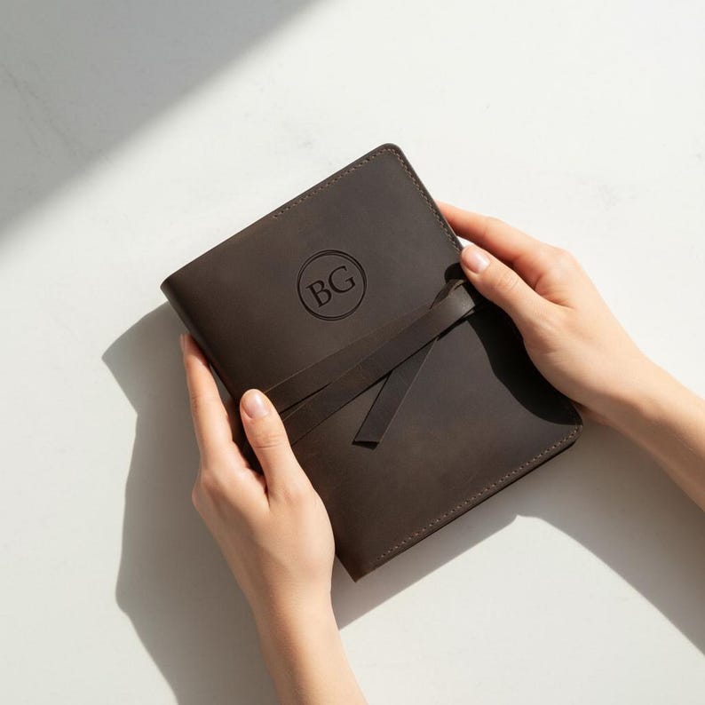 May include: A dark brown leather journal with a wrap-around strap. The journal has a circular monogram with the letters "BG" embossed on the cover. The journal is held open by two hands.